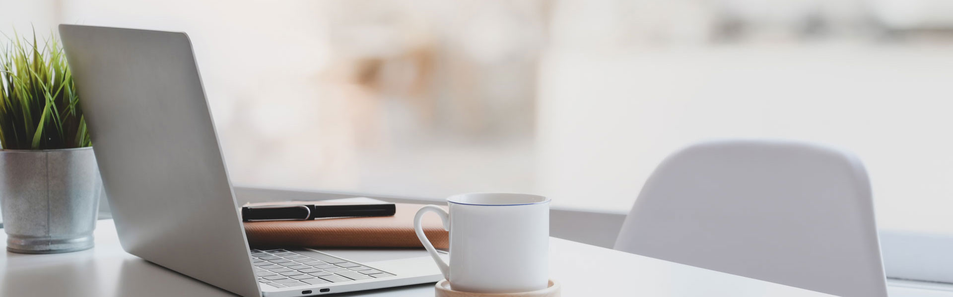 Schreibtisch mit aufgeklappten Computer, Pflanze, Tasse und leerem Stuhl